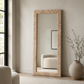A tall floor mirror with a woven natural-fiber frame stands in a neutral, modern room next to a window, a beige armchair, and a large ceramic vase with dried branches.