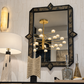 An ornate rectangular wall mirror with a black and gold patterned frame, mounted on a textured white wall above a wooden console table. A matching gold multi-arm chandelier is reflected in the mirror, alongside a table lamp, decorative sculpture, and glass decanters.