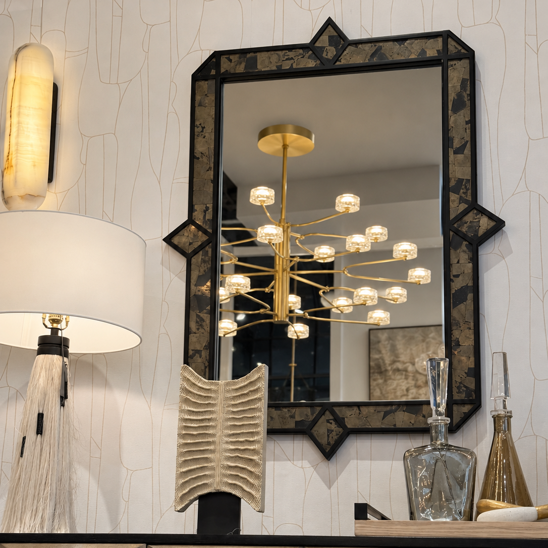 An ornate rectangular wall mirror with a black and gold patterned frame, mounted on a textured white wall above a wooden console table. A matching gold multi-arm chandelier is reflected in the mirror, alongside a table lamp, decorative sculpture, and glass decanters.