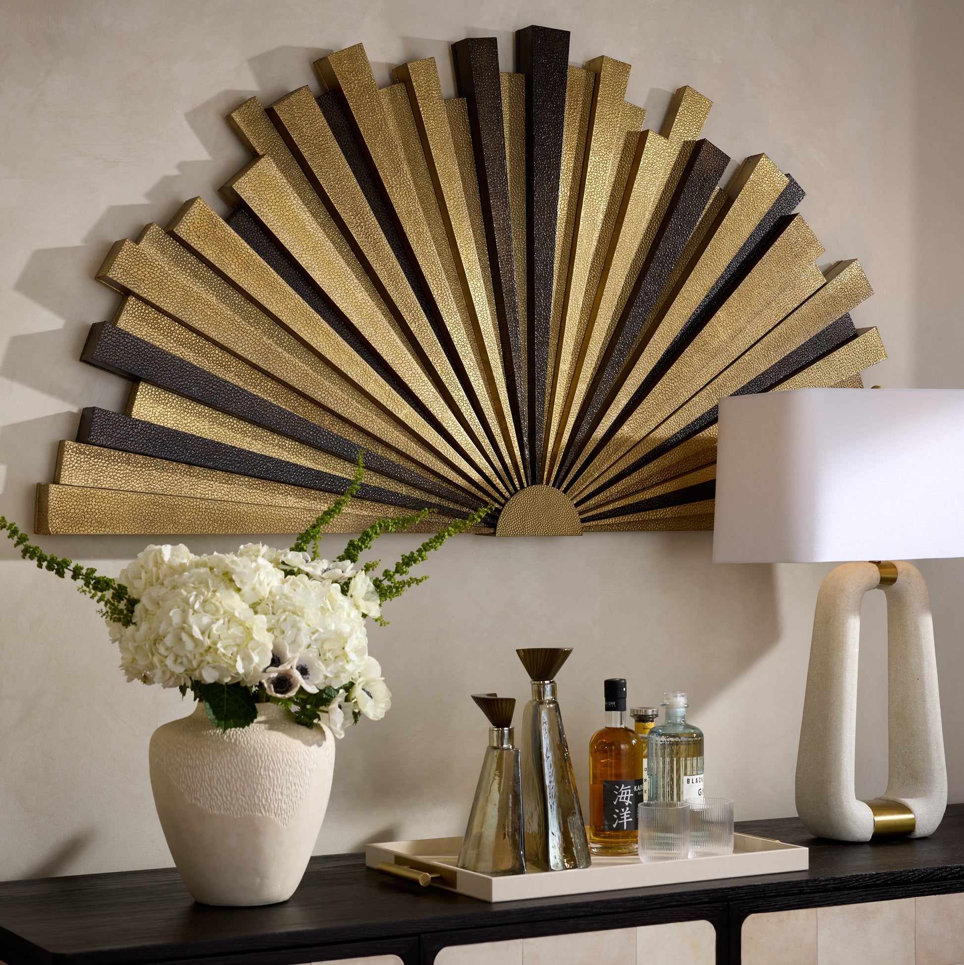 Decorative wall art with a sunburst design above a console table with a lamp, bottles, and flowers.
