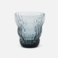 A ribbed, fluted glass tumbler with a gradient color transition from light blue-gray at the top to a deep black at the base, shown on a clean white background.