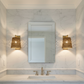 A modern bathroom vanity featuring a rectangular wall mirror, a white marble countertop with an integrated sink, and two matching gold sconces mounted on a white marble wall.