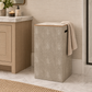 A beige fabric laundry hamper with a wood-look top trim and a draped cream towel, set in a modern bathroom next to a vanity.
