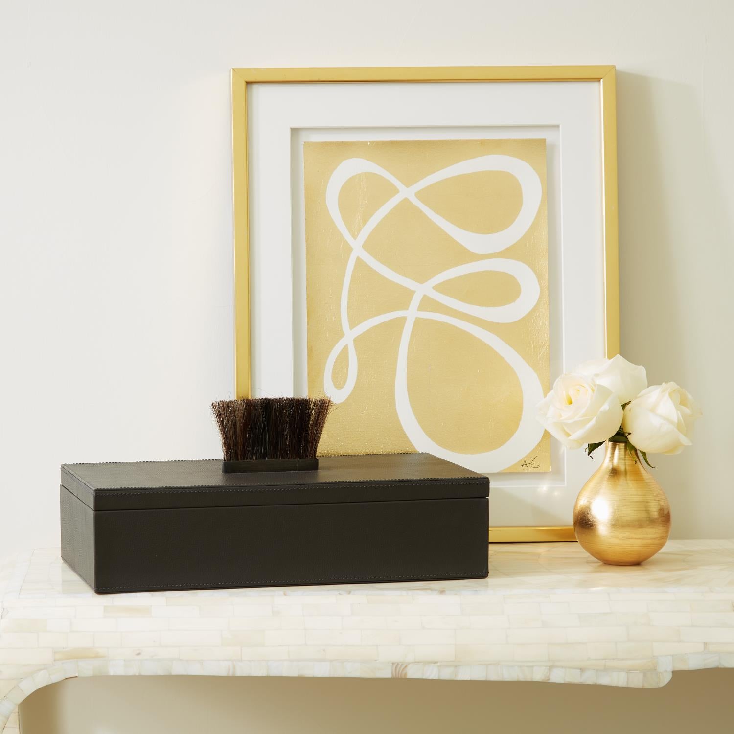 Decorative setup with a black box, brush, gold vase with flowers, and abstract art on a white surface.