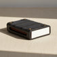 A black textured leather card holder case with brass-tone rivets and a white front label area, photographed on a clean light gray background.