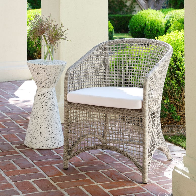 Wicker chair with a white cushion on a patio with a small table and plants.