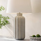 Textured table lamp with a white shade on a surface next to a plant and bowl of fruit.