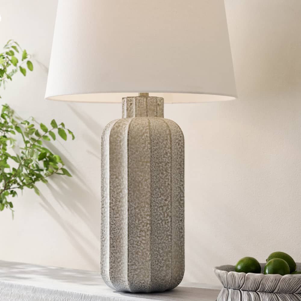 Textured table lamp with a white shade on a surface next to a plant and bowl of fruit.