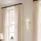 A pair of long cream pleated curtains hanging from a black rod over a white window in a bright room with white paneled walls and a wall-mounted light.