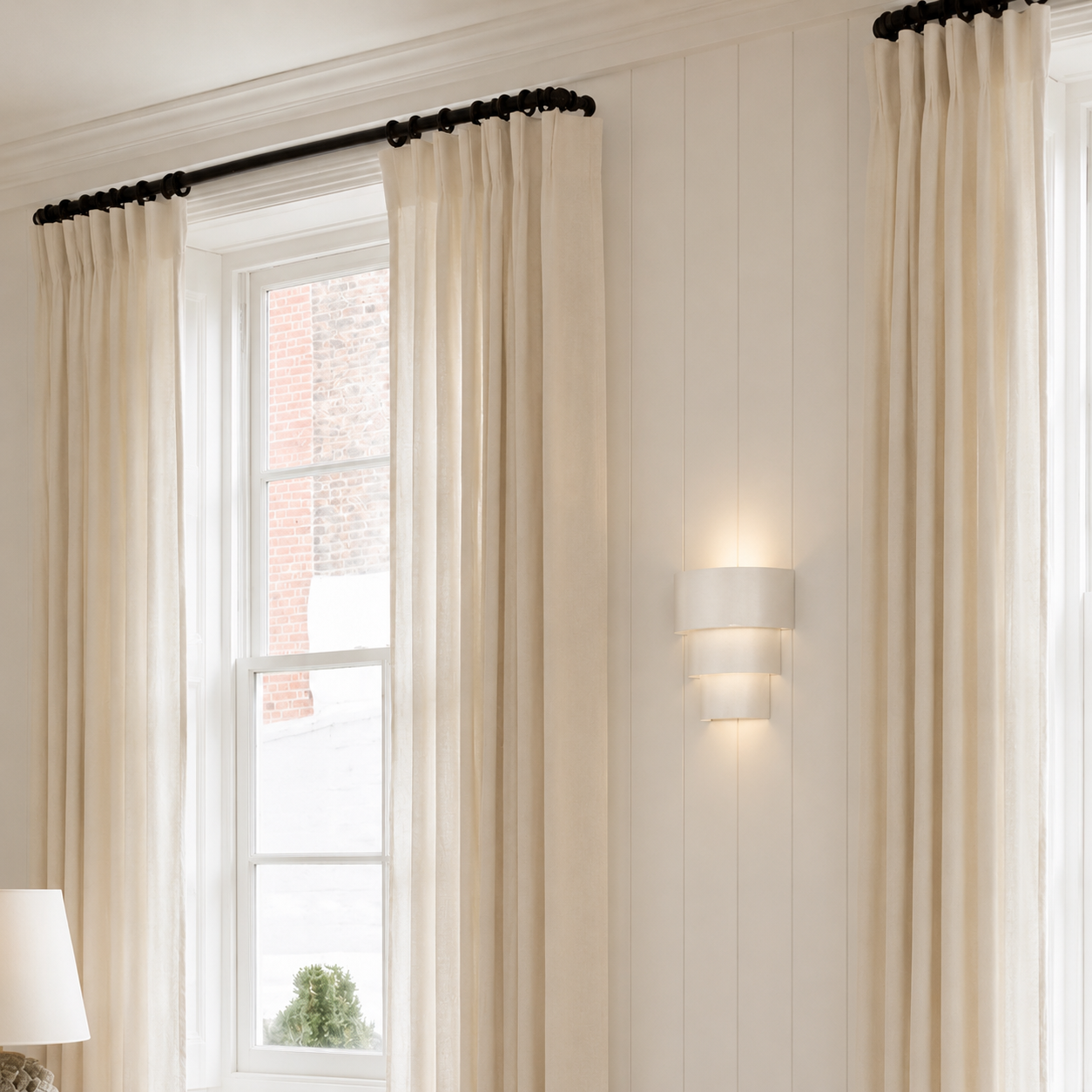 A pair of long cream pleated curtains hanging from a black rod over a white window in a bright room with white paneled walls and a wall-mounted light.