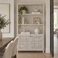 A tall white wooden bookcase with three open shelves and two lower cabinet doors with woven cane panels, styled with books, vases, and a hanging plant in a bright dining room.
