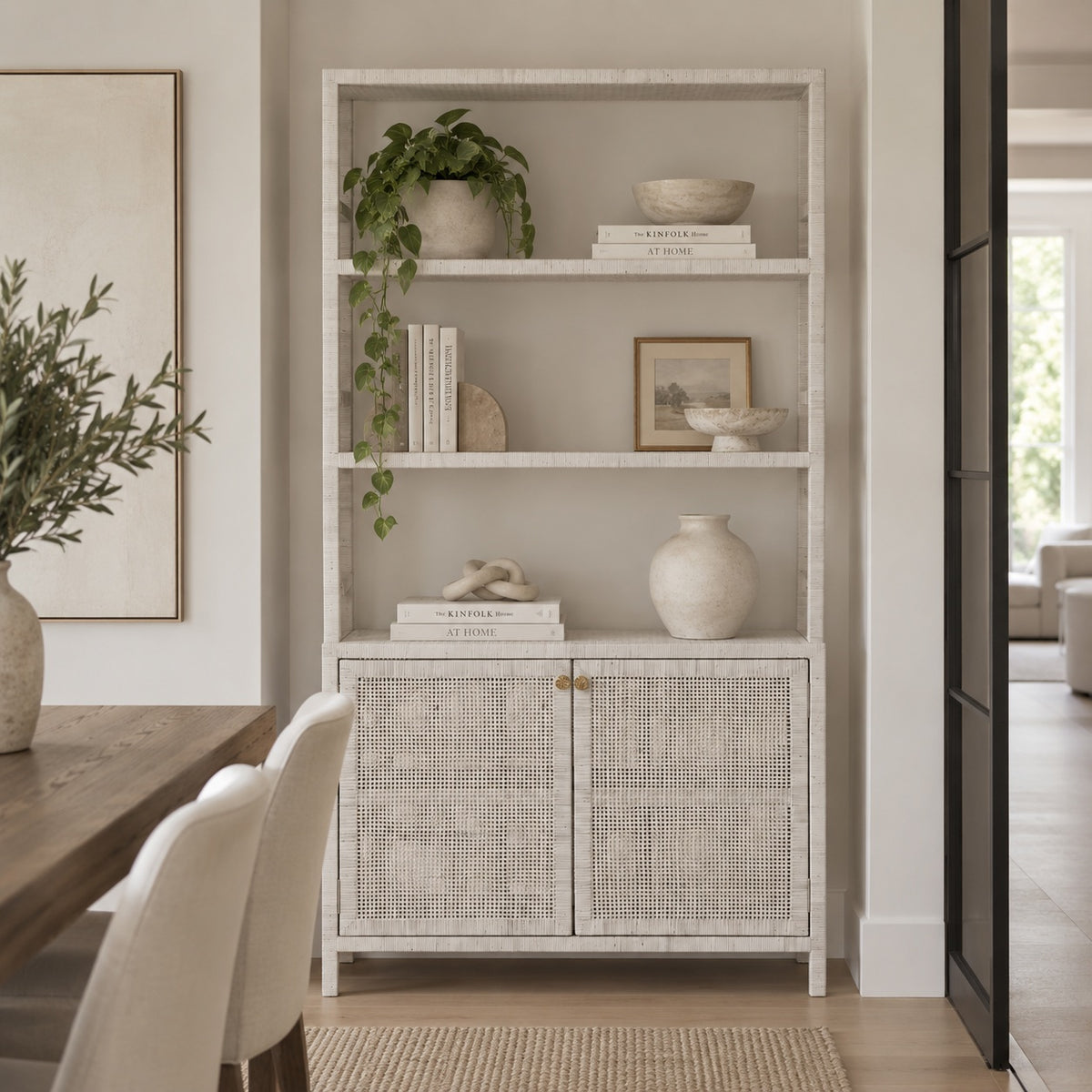 A tall white wooden bookcase with three open shelves and two lower cabinet doors with woven cane panels, styled with books, vases, and a hanging plant in a bright dining room.