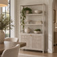 A tall, multi-tiered open shelving cabinet with a distressed white finish and woven cane doors, styled with home decor in a bright, modern dining room.