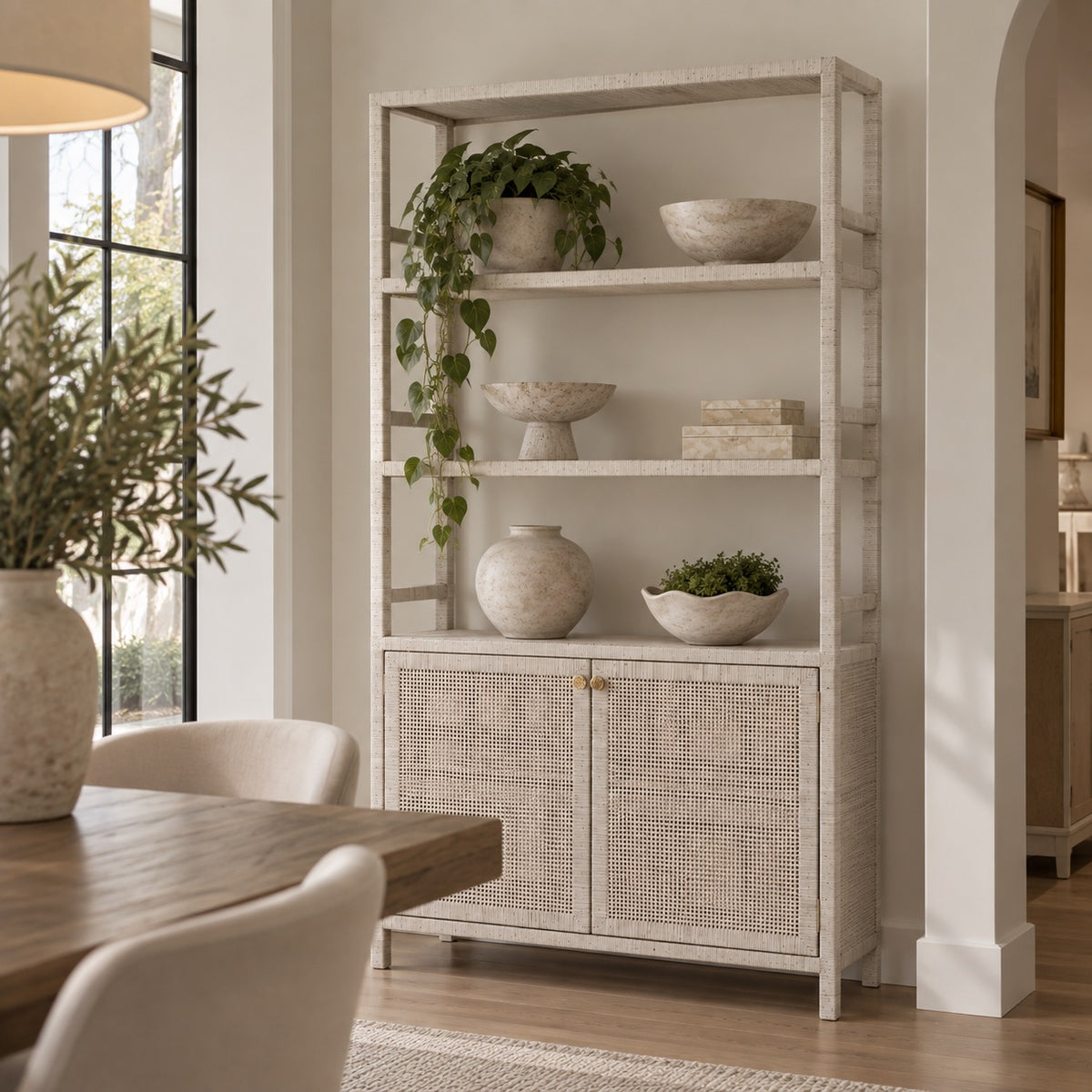 A tall, multi-tiered open shelving cabinet with a distressed white finish and woven cane doors, styled with home decor in a bright, modern dining room.