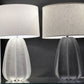 Two modern table lamps with textured drum shades and sculptural tulip-shaped bases, shown side by side against a dark gray background.