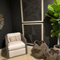 A modern accent chair with white cushions and a gray woven frame, styled in a room setting with a striped pillow, decorative baskets, a potted fiddle-leaf fig plant, and black geometric wall art.