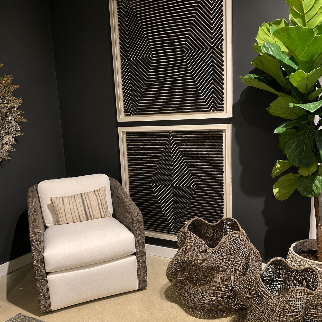 A modern accent chair with white cushions and a gray woven frame, styled in a room setting with a striped pillow, decorative baskets, a potted fiddle-leaf fig plant, and black geometric wall art.