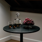Small round table with a bottle, two glasses, and a bowl of grapes under a skylight.