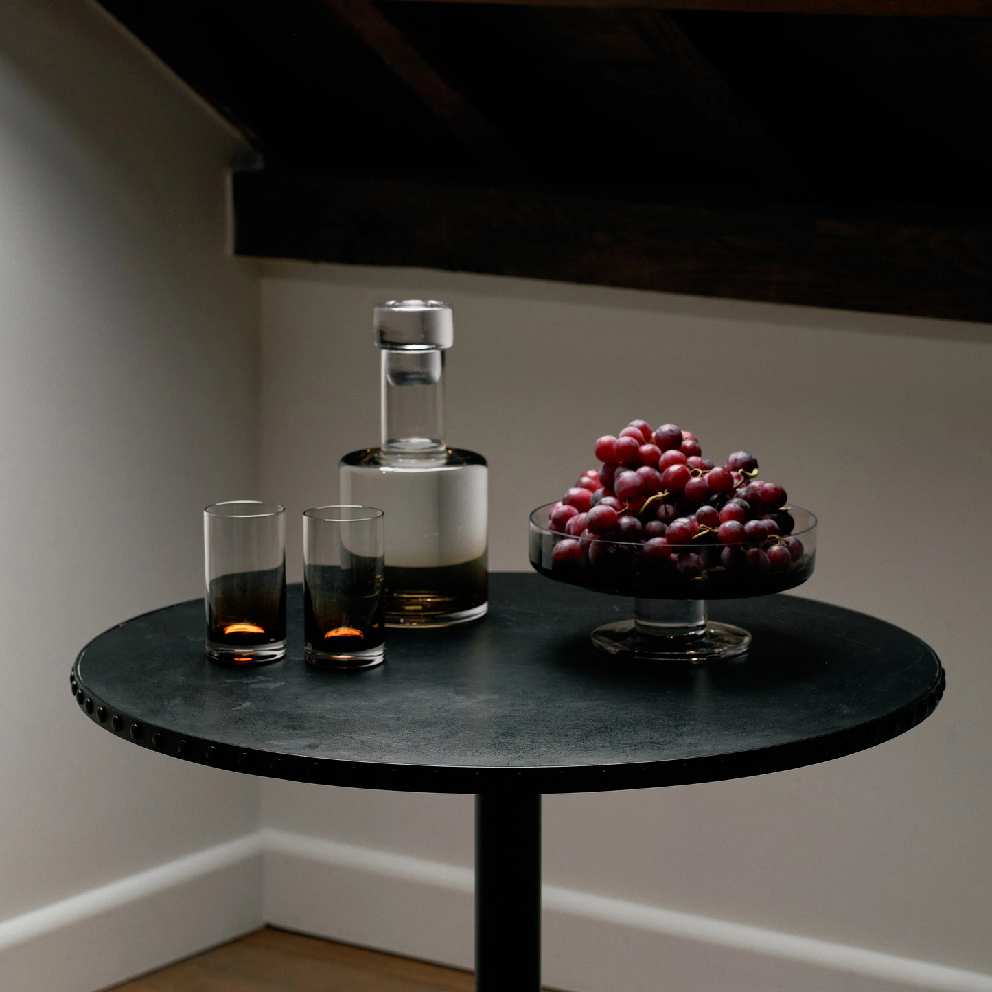 Small round table with a bottle, two glasses, and a bowl of grapes under a skylight.