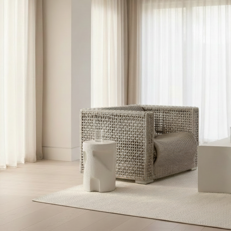 A modern woven armchair in a neutral beige tone, styled in a minimalist, bright room with sheer curtains, a low white side table, and a textured rug.