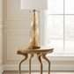 Decorative table lamp with gold base and white shade on a wooden side table in a room with large windows.