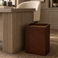 A dark brown woven basket-style trash can with a slanted open top, placed on a textured beige rug next to a modern wood desk in a warm, neutral-toned room.