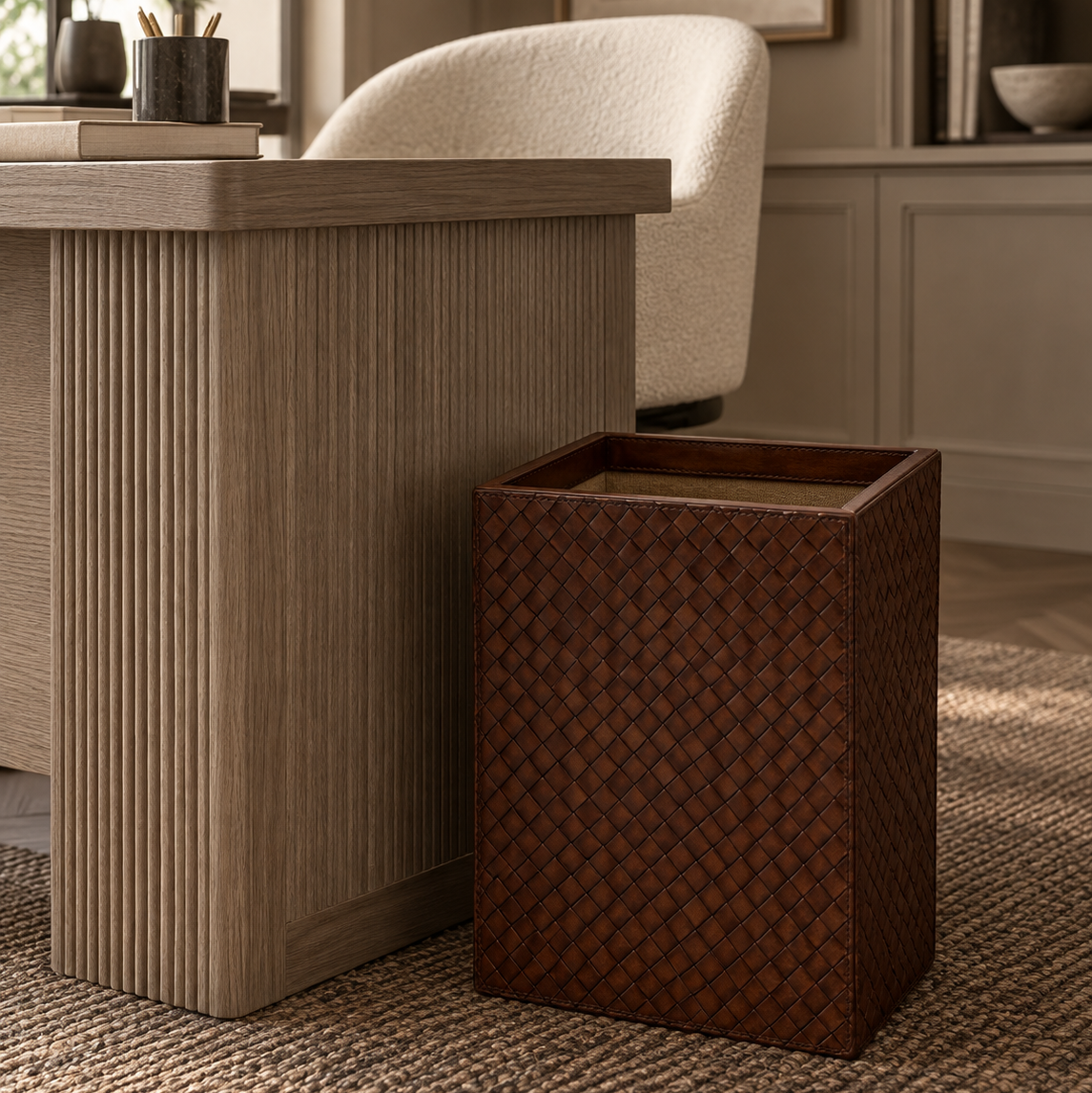 A dark brown woven basket-style trash can with a slanted open top, placed on a textured beige rug next to a modern wood desk in a warm, neutral-toned room.