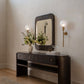 A dark wood console table with a matching rectangular mirror, styled in a bright entryway with a wall sconce, floral arrangement, and an arched wooden door in the background.