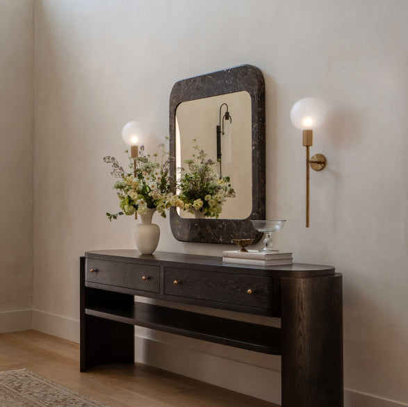 A dark wood console table with a matching rectangular mirror, styled in a bright entryway with a wall sconce, floral arrangement, and an arched wooden door in the background.