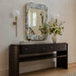 A dark wood console table with drawers and an open shelf, styled in a home entryway setting under a patterned rectangular mirror, with a vase of flowers and wall sconces.