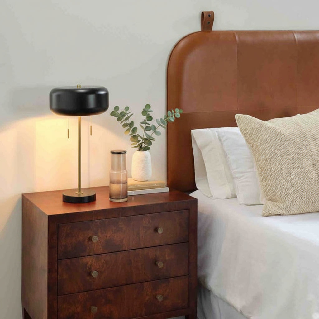 Wooden nightstand with lamp and decor next to a bed with a brown leather headboard.