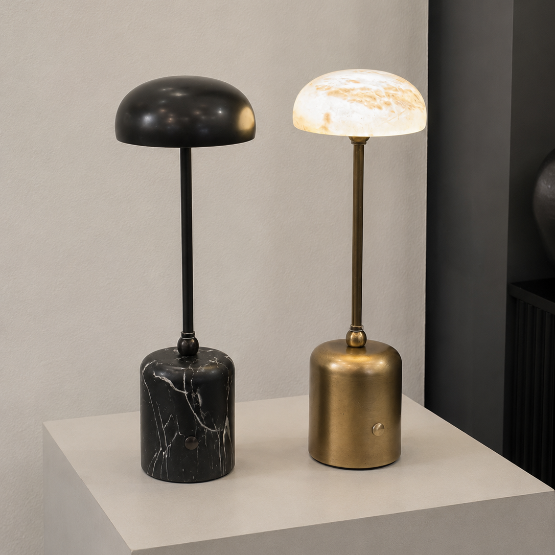 Two modern table lamps displayed on a white pedestal against a neutral gray wall. The left lamp has a black dome shade and a black marble base.