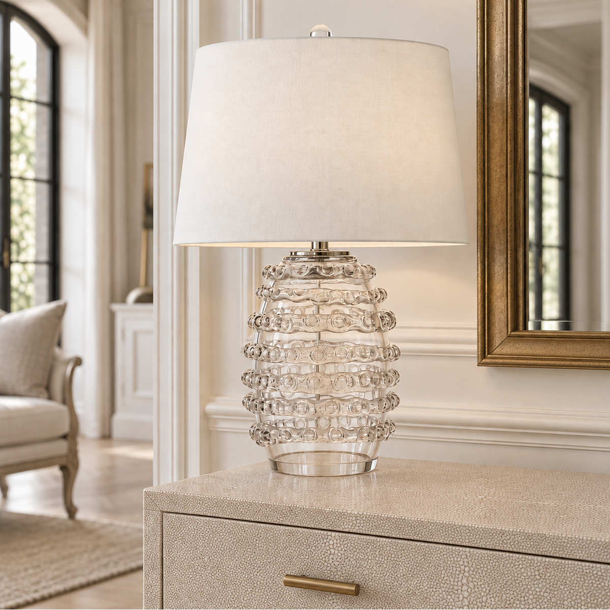 A table lamp with a textured clear glass base and a large off-white fabric drum shade, styled on a light-colored textured dresser in a bright, elegant room.