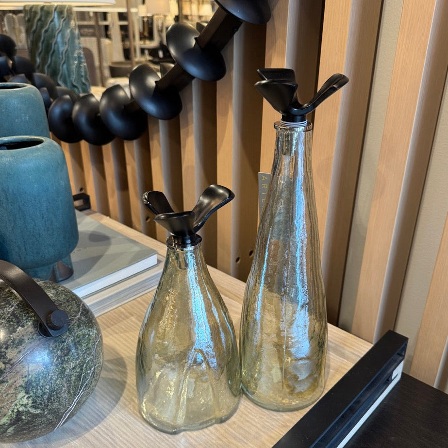 Two glass bottles with black nozzles on a wooden surface, with a blurred background of indoor decor.