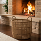 A natural woven rattan laundry basket with two loop handles sits on a dark wood floor in front of a warm stone fireplace with a fire burning inside.