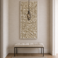 A large vertical textured wall art piece in warm beige and cream tones hangs on a light beige wall above a tufted bench in a bright entryway. A hanging pendant light is suspended in front of the artwork.