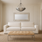 A modern living room scene featuring a cream two-seater sofa, a matching tufted ottoman with a chrome metal frame, a decorative rectangular mirror, and a tiered metal pendant chandelier.