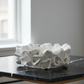 A white, textured, ruffled ceramic bowl or planter displayed on a black marble tabletop in a modern room setting.