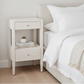 A light beige wooden nightstand with two drawers and open shelving, styled beside a bed with white bedding and pillows in a bright bedroom.