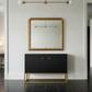 A black two-door cabinet with a gold metal frame base and small gold square handles, positioned beneath a large ornate gold mirror in a white hallway.