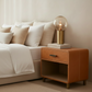 A modern tan leather nightstand beside a bed with white linens and beige pillows, holding a brass table lamp with a clear glass globe bulb and stacked books.