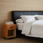 A modern bedroom scene featuring a bed with crisp white linens, a black leather headboard, a warm brown wooden nightstand with books, and a textured beige woven wall background.