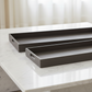 Two dark gray textured rectangular serving trays with cutout handles, displayed on a white marble table with a soft-focus background of curtains.