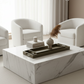 A modern square coffee table with a white marble pattern, styled in a bright living room with two white armchairs and a dark tray holding books and decorative objects.