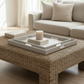 A square woven rattan coffee table in a bright living room, styled with a white tray, books, and a candle, with a beige sofa in the background.