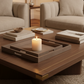 A styled living room scene with a wooden coffee table in the foreground, holding a gray textured tray with a lit white candle, stacked books, and small decor items. Two upholstered armchairs, a floor lamp, and a fireplace are visible in the background.