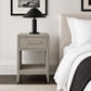 A modern bedside table in a warm gray textured finish, styled in a bedroom next to a bed with white linens. A black table lamp with a gold handle sits on top.