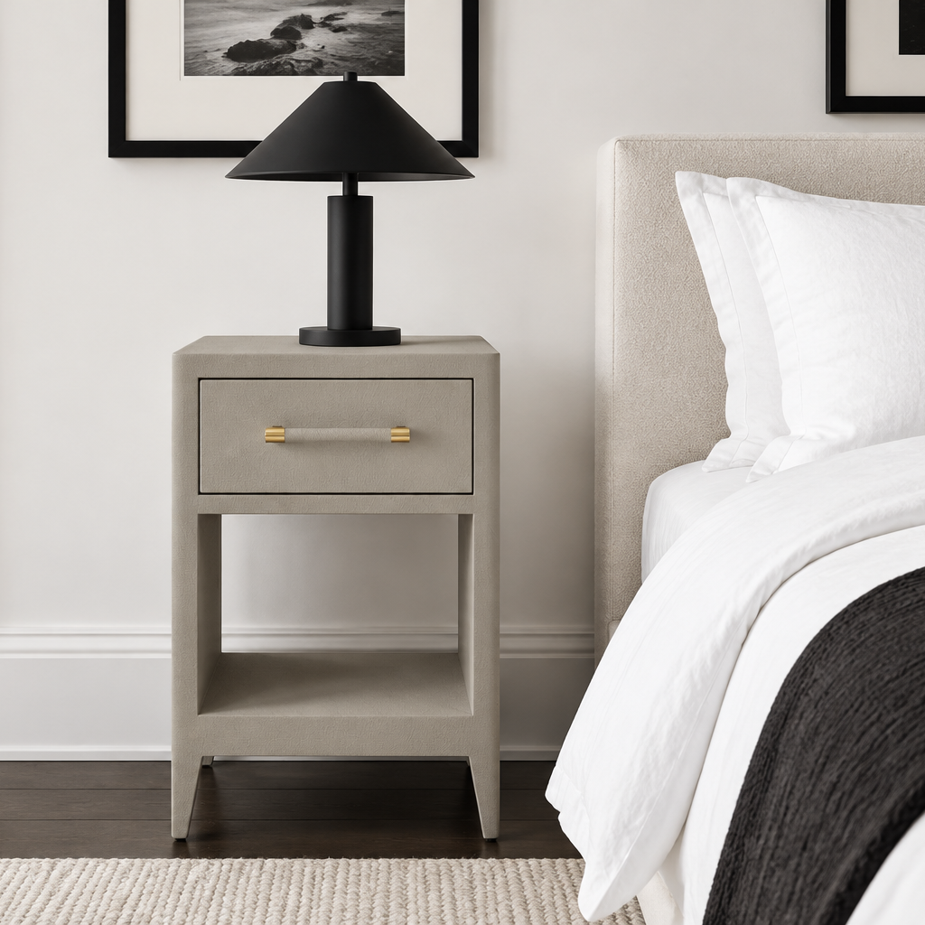 A modern bedside table in a warm gray textured finish, styled in a bedroom next to a bed with white linens. A black table lamp with a gold handle sits on top.
