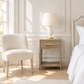 A styled bedroom corner featuring a cream upholstered accent chair next to a light wood nightstand with a white table lamp, set against a beige paneled wall with a bed partially visible on the right.
