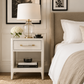 A white nightstand in a styled bedroom setting, featuring a clear glass lamp base with a white drum shade, a vase of white flowers, and decorative books on the shelf.